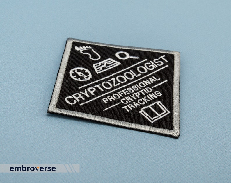May include: Black and white embroidered patch with the text "Cryptozoologist Professional Cryptid Tracking" and various symbols including a compass, footprint, magnifying glass, and an open book.