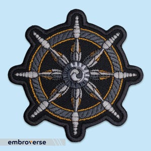 Könnte beinhalten: Gestickter Aufnäher mit einem Steuerraddesign. Der Aufnäher hat einen schwarzen Hintergrund mit grauen und goldenen Akzenten. Die Speichen des Rades sind grau, und der äußere Rand ist goldfarben umrandet. Das Wort "embroverse" steht unten.