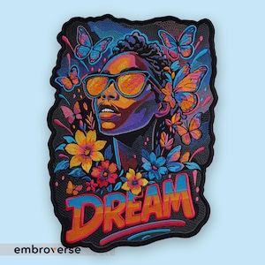 Pode incluir: Patch bordado com um retrato colorido de uma pessoa usando óculos de sol, rodeada por flores e borboletas. A palavra "DREAM" é exibida em letras multicoloridas em negrito. O patch tem uma borda preta.