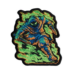 Puede incluir: Un parche bordado que muestra una figura encapuchada en azul y naranja, empuñando dos hojas. La figura está rodeada de humo verde arremolinado, todo delineado con hilo negro sobre un fondo oscuro, creando un diseño gráfico detallado.