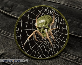 Puede incluir: Araña verde bordada en un parche de tela negro con una telaraña blanca. El parche tiene un borde verde y es circular.