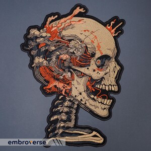 May include: Embroidered patch featuring a detailed skull design with exposed internal structures and fiery accents. The patch has a beige background with orange, gray, and black detailing. The word "embroverse" is printed at the bottom.