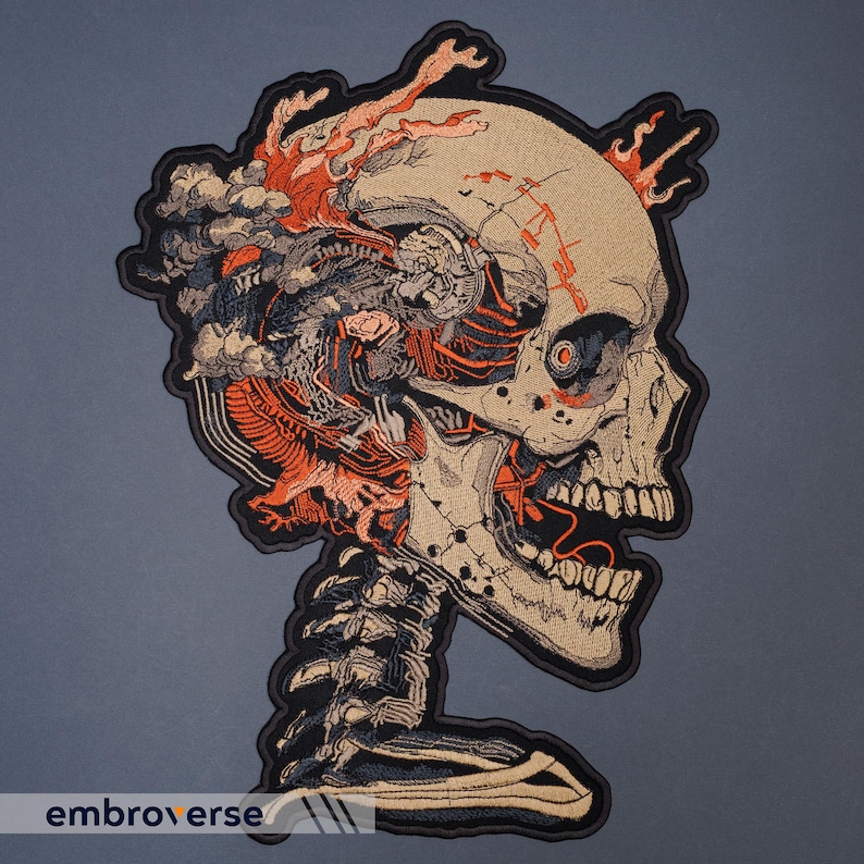 May include: Embroidered patch featuring a detailed skull design with exposed internal structures and fiery accents. The patch has a beige background with orange, gray, and black detailing. The word "embroverse" is printed at the bottom.
