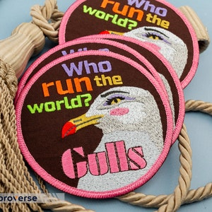 May include: Three brown embroidered patches with pink stitching. Each patch features a white seagull with the text "Who run the world? Gulls" in colourful letters.
