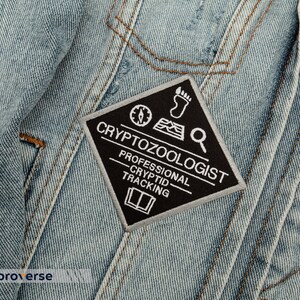 May include: A black and white embroidered patch with the text "Cryptozoologist - Professional Cryptid Tracking" on a diamond-shaped background. The patch features a compass, footprint, and magnifying glass.