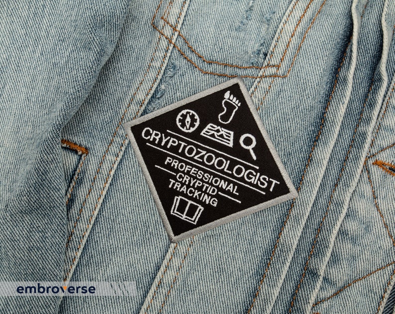 May include: A black and white embroidered patch with the text "Cryptozoologist - Professional Cryptid Tracking" on a diamond-shaped background. The patch features a compass, footprint, and magnifying glass.