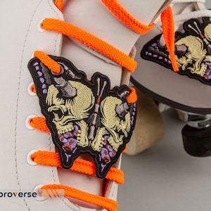 Puede incluir: Primer plano de patines blancos con cordones naranja brillante. Un parche negro con un diseño de calavera está adherido a los cordones. El parche tiene bordados amarillos, morados y grises. La palabra "embroverse" está impresa en blanco en la parte inferior.