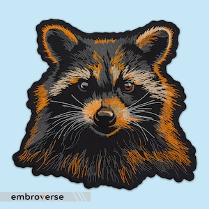 May include: Embroidered patch of a raccoon's face with black fur, orange and grey markings, and a white nose.