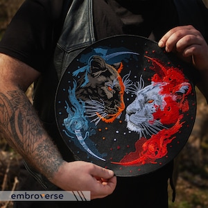 Peut inclure: Un patch brodé noir et blanc avec deux visages d'animaux. Le côté gauche présente une panthère noire avec des flammes bleues et blanches. Le côté droit présente un lion blanc avec des flammes rouges et orange. Le patch est circulaire et a un fond noir.
