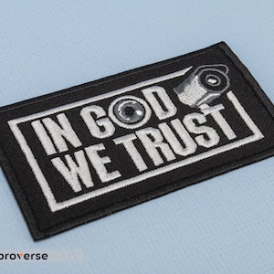 May include: Black embroidered patch with white text that reads "In God We Trust" and a stylised eye with a security camera above it.