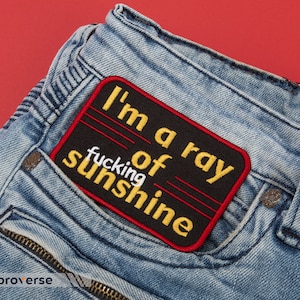 Peut inclure: Un écusson brodé noir et rouge avec du texte jaune qui dit "I'm a ray of fucking sunshine" sur un jean bleu.
