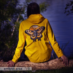 Könnte beinhalten: Ein gelber Regenmantel mit einem großen gestickten Schmetterling auf der Rückseite. Der Schmetterling ist dunkelbraun und orangefarben mit schwarzen Akzenten.