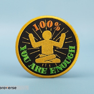 Op de afbeelding: Een geborduurde patch in zwart en geel met een persoon in een yogapose en de tekst "100% You Are Enough".