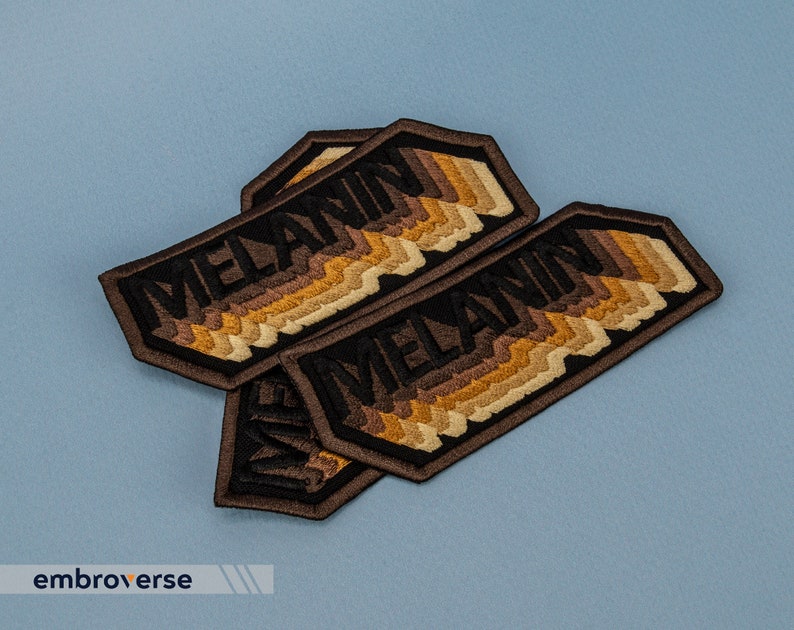Peut inclure: Trois patchs brod&eacute;s marron et noir avec le mot "MELANIN" en lettres noires. Les patchs ont un design en couches avec des nuances de marron.