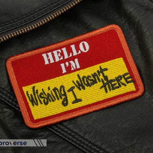 May include: Red and yellow embroidered patch with the text "Hello I'm Wishing I Wasn't There" on a black leather jacket.