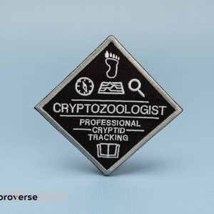May include: Black and white embroidered patch in a diamond shape. The patch reads "Cryptozoologist - Professional Cryptid Tracking" and features a compass, footprint, map, and magnifying glass.
