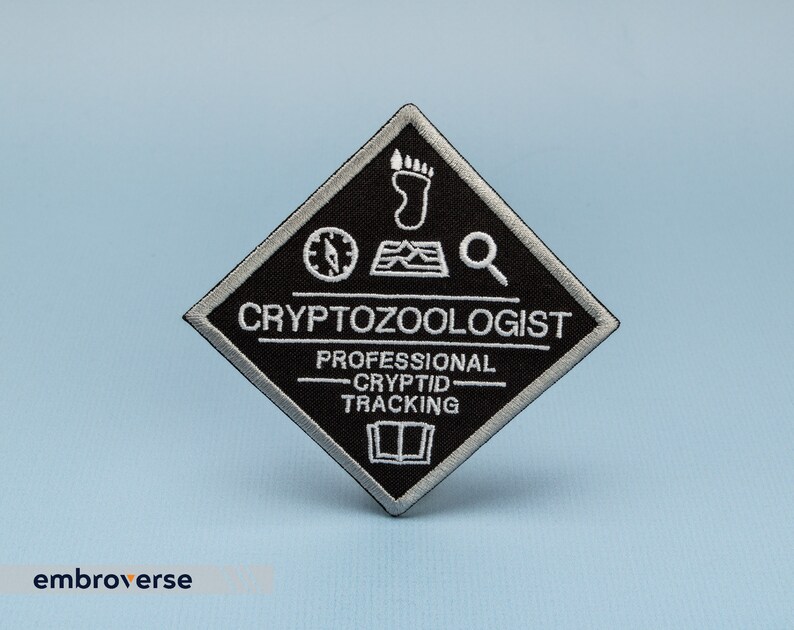 May include: Black and white embroidered patch in a diamond shape. The patch reads "Cryptozoologist - Professional Cryptid Tracking" and features a compass, footprint, map, and magnifying glass.