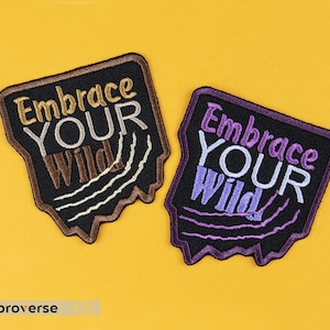 May include: Two embroidered patches with a black background and a white outline. The patches are shaped like a dripping candle. The text on the patches reads "Embrace Your Wild". One patch is brown with gold text and the other is purple with white text.