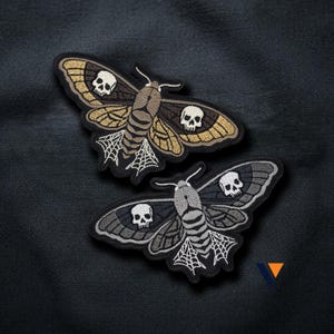 以下が含まれることがあります： 羽にドクロのモチーフが刺繍された2枚の蛾のワッペン。上のワッペンはベージュと茶色の羽に白いドクロ、下のワッペンはグレーと黒の羽に白いドクロが描かれており、どちらも白いクモの巣のような模様が施されています。