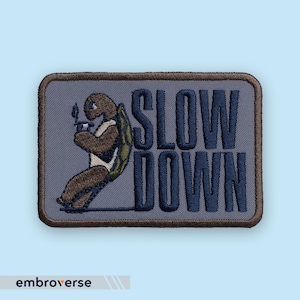 Könnte beinhalten: Gestickter Aufnäher mit einer Zeichentrickschildkröte, die eine Pfeife raucht, mit den Worten "SLOW DOWN" in Marineblau. Der Aufnäher hat einen grauen Hintergrund und einen braunen Rand. Die Schildkröte hat einen grünen Panzer.