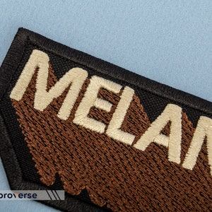 Peut inclure: Un patch noir avec des coutures brunes et le mot "MELANIN" brod&eacute; en blanc.