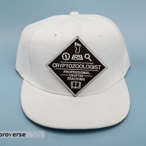 May include: White baseball cap with a black and grey patch that reads "Cryptozoologist - Professional Cryptid Tracking". The patch features a compass, footprint, and magnifying glass.