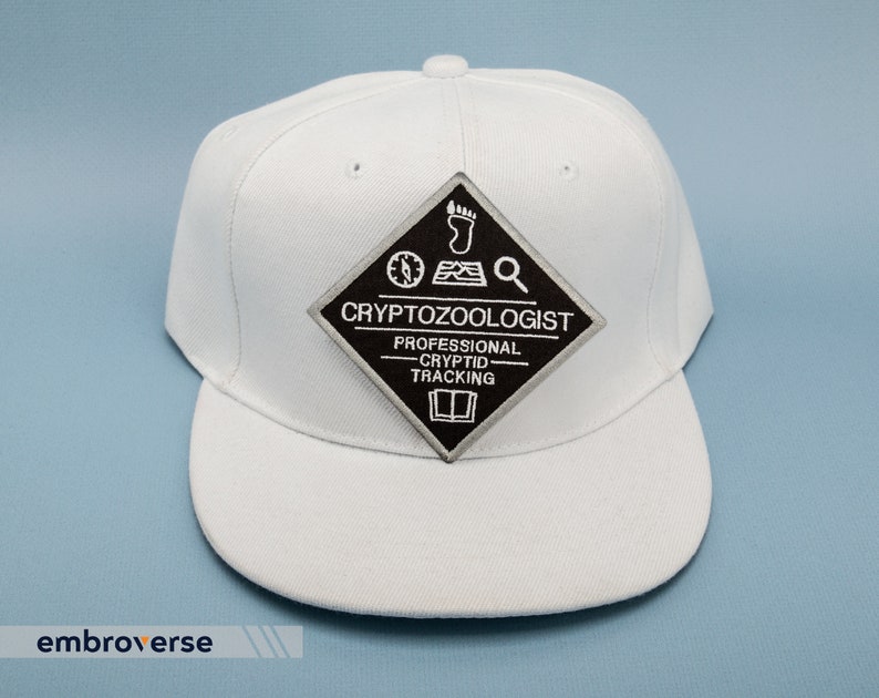 May include: White baseball cap with a black and grey patch that reads "Cryptozoologist - Professional Cryptid Tracking". The patch features a compass, footprint, and magnifying glass.