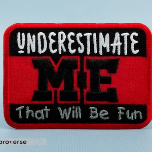 May include: Red embroidered patch with white text that reads "UNDERESTIMATE ME That Will Be Fun" in a black outlined font.