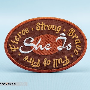 May include: An oval-shaped embroidered patch in shades of brown and orange. The patch features the words "Fierce, Strong, Brave" and "Full of Fire" in a circular pattern around the edge. The words "She Is" are in the center.