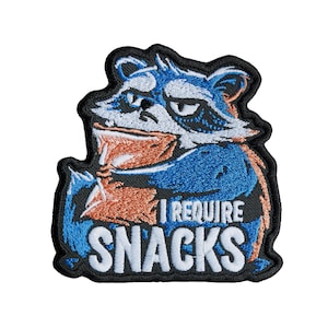 May include: An embroidered patch featuring a blue and white raccoon with a grumpy expression, holding an orange snack bag. The text "I REQUIRE SNACKS" is stitched in white capital letters at the bottom against a black border outline.
