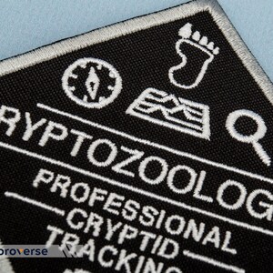 May include: White embroidered patch on black fabric with the text "CRYPTOZOOLOGIS PROFESSIONAL CRYPTID TRACKING" and icons of a compass, footprint, and magnifying glass.