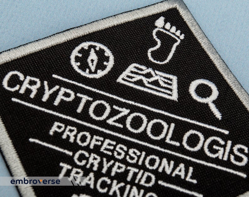 May include: White embroidered patch on black fabric with the text "CRYPTOZOOLOGIS PROFESSIONAL CRYPTID TRACKING" and icons of a compass, footprint, and magnifying glass.