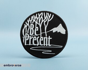 Może przedstawiać: Czarna naszywka haftowana z białym tekstem "Be Present" i stylizowanym wzorem drzewa.