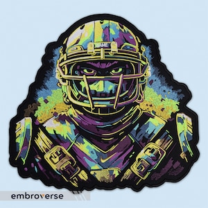 Op de afbeelding: Een geborduurde patch van een American footballspeler die een helm draagt met het woord "GAME" erop. De speler draagt een paars en groen uniform met gouden accenten. De patch is op een zwarte achtergrond.