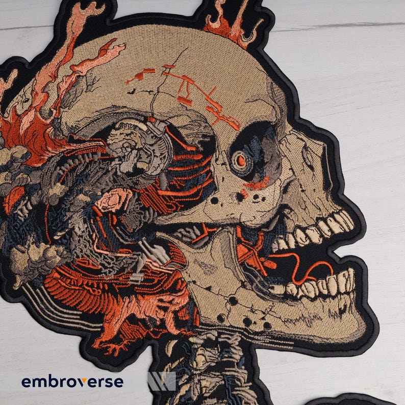 May include: Embroidered patch featuring a detailed, stylized skull with exposed internal components. The design incorporates shades of tan, black, and orange, with intricate detailing and a black border. The patch has the word "embroverse" printed on it.