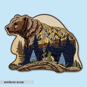 Pode incluir: Etiqueta bordada de um urso castanho em pé em frente a uma cordilheira com uma floresta em primeiro plano. O urso é delineado em castanho e as montanhas estão em tons de amarelo, laranja e azul.