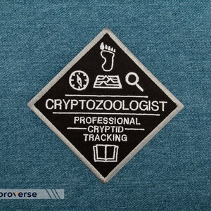 May include: Black and white embroidered patch in a diamond shape. The patch features a compass, footprint, magnifying glass, and book icon. The text reads "Cryptozoologist Professional Cryptid Tracking".