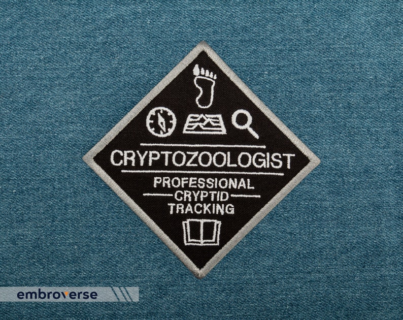 May include: Black and white embroidered patch in a diamond shape. The patch features a compass, footprint, magnifying glass, and book icon. The text reads "Cryptozoologist Professional Cryptid Tracking".