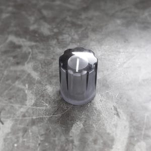 May include: A clear, cylindrical control knob with a scalloped top and a white vertical line. The knob is made of a translucent material and sits on a gray surface. The design is simple and modern.