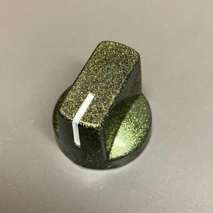 May include: A cylindrical control knob with a dark olive green finish and a fine glitter texture. The knob features a flat top surface with a single vertical white indicator line running down the side to show the current position setting.