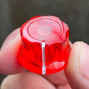 May include: A translucent red plastic control knob with a circular top and a fluted side profile. A single vertical white line is printed on the side of the knob to indicate position. The knob has a hollow center and a smooth, glossy finish.