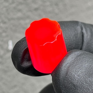 Puede incluir: Una pequeña tapa cilíndrica de color rojo vibrante con un borde superior ondulado y festoneado. El objeto tiene un acabado liso de color sólido y una forma redondeada distintiva con un borde texturizado, presentado como un componente único.