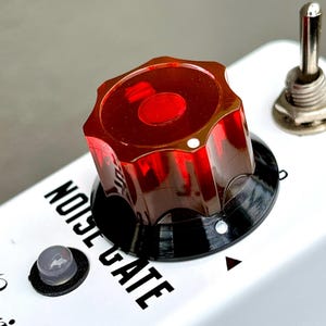 May include: A close-up of a white electronic device with a red, translucent knob and the words "NOISE GATE" printed in black. A small, clear button and a silver toggle switch are also visible.