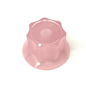 Op de afbeelding: Een doorschijnende roze knop met een stervormige bovenkant en een cirkelvormige basis. De knop heeft een reeks afgeronde randen en twee kleine gaatjes aan de basis. Het object staat tegen een witte achtergrond.