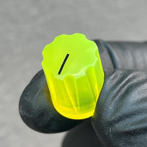 May include: A neon green control knob with a black line indicator. The knob has a ridged edge and is held by a gloved hand. The knob is a bright, translucent green color.