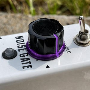 May include: A white guitar effects pedal labeled NOISE GATE with a black control knob featuring a purple base ring. The pedal includes a silver metal toggle switch, a small circular indicator light, and a metal input jack on the front edge.