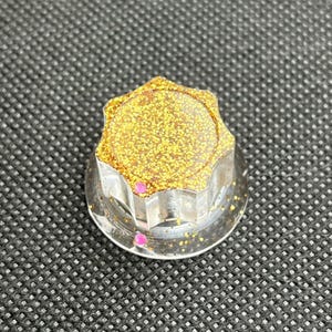 May include: A clear, faceted knob with a gold glitter top and small pink accents. The knob is likely for a piece of furniture or equipment, adding a decorative touch. The design is modern and eye-catching.