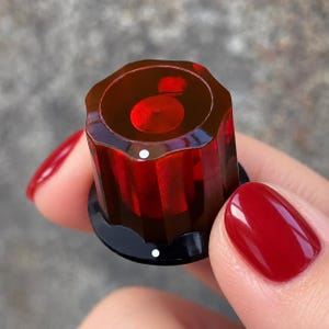 May include: A cylindrical control knob featuring a translucent deep red fluted body with a smooth top surface. The knob includes a black circular base and two small white indicator dots, one on the top edge and one on the black base.