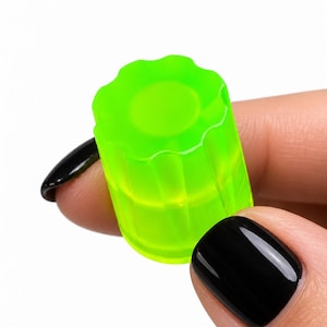 May include: A small, translucent, neon green cylindrical cap with a fluted, gear-like top edge. The object features a smooth, semi-transparent finish and a distinct ridged design around its upper circumference, creating a textured appearance.