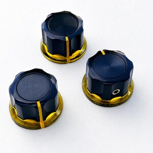 May include: Three circular control knobs featuring a black fluted exterior with a gold-coloured base and a vertical yellow indicator line. Each knob has a flat top surface and a small metal set screw visible on the side for secure attachment.
